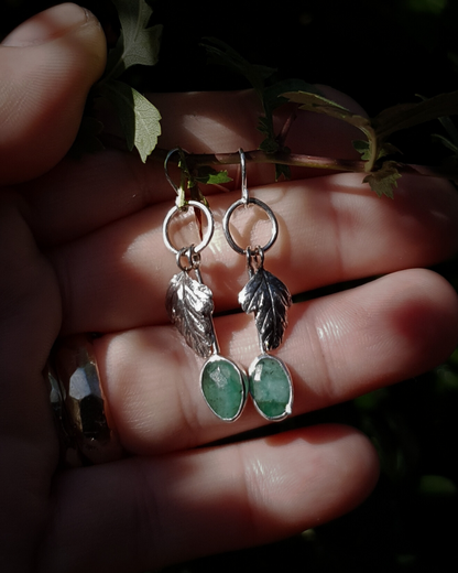 Two silver hook earrings sit in an open palm to demonstrate their size. They are comprised of a little silver circlet from which hangs a solid silver leaf. A faceted emerald also hangs from the circlet at the end of a long stem so it hangs below the leaf. The emeralds are rose-cut and rather unrefined showing different shades of green and the leaves are complete with natural details including veins. The earrings are illuminated in natural sunlight.