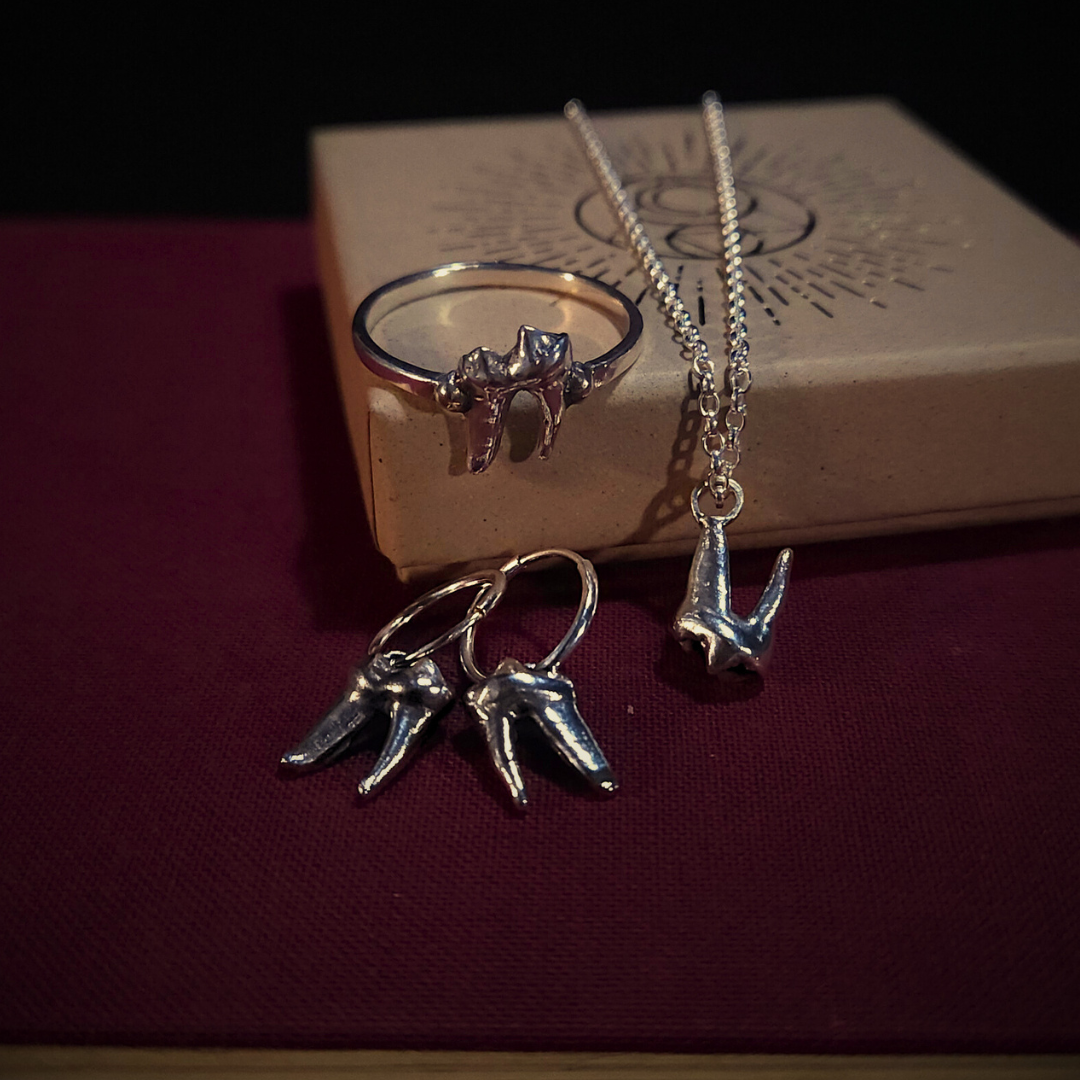 A set of sterling silver fox tooth jewellery sit atop a cloth-bound red book and next to a natural cardboard jewellery box. The set is comprised of a ring, earrings and a necklace.