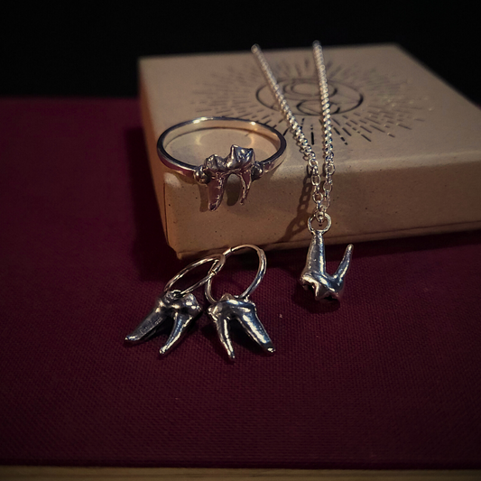 A set of sterling silver fox tooth jewellery sit atop a cloth-bound red book and next to a natural cardboard jewellery box. The set is comprised of a ring, earrings and a necklace.