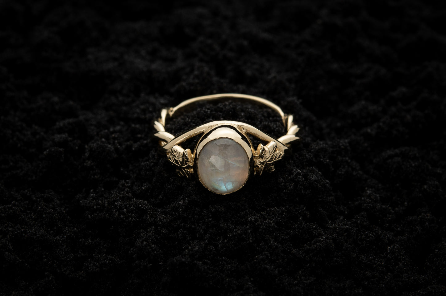 A botanical engagement ring made from rainbow moonstone and 9 carat yellow gold lays atop black sand. It features a twisted band and an ivy leaf on each shoulder. The moonstone shines gently in the camera flash, teasing it's bluey rainbow hues.