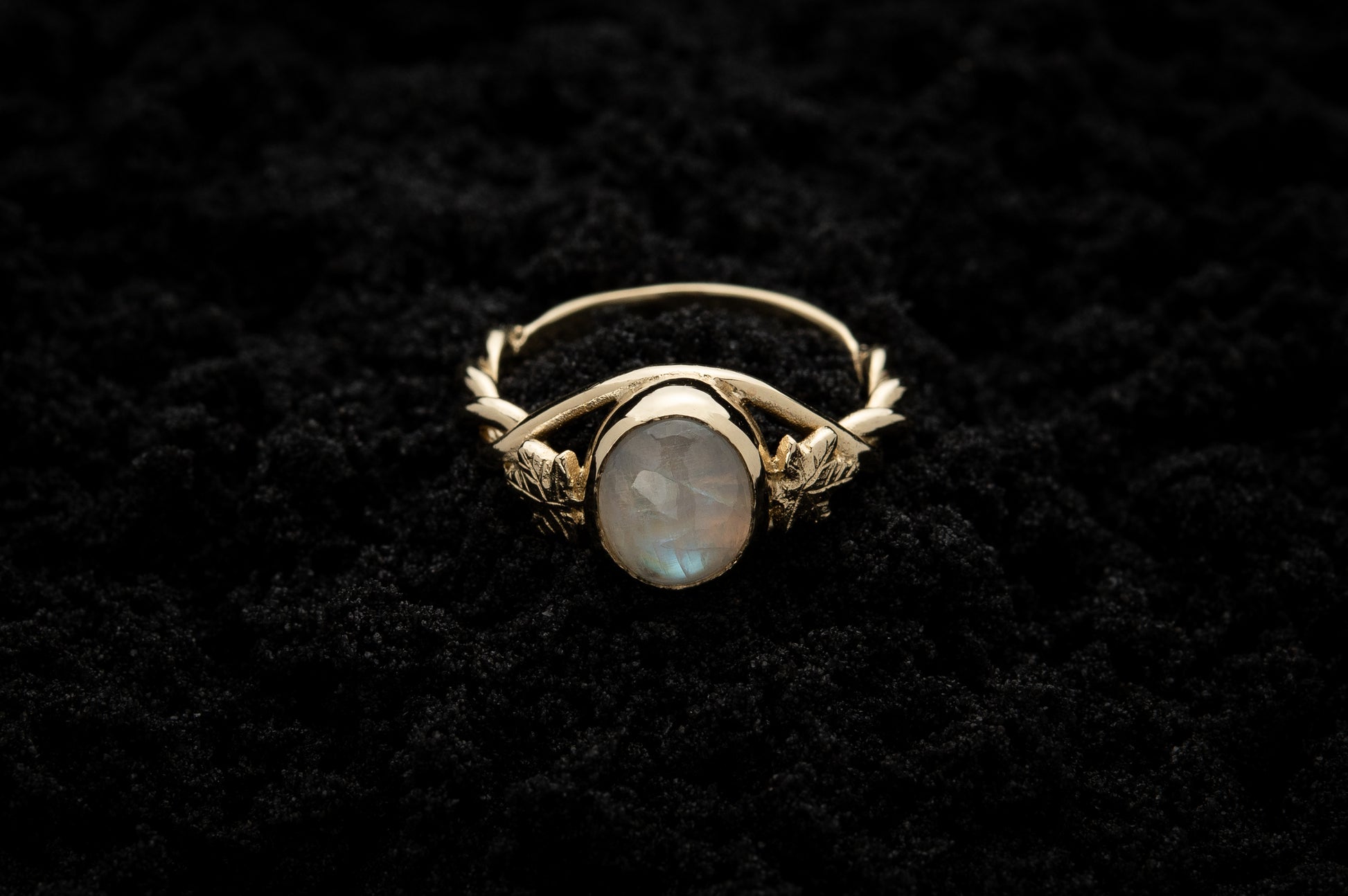A botanical engagement ring made from rainbow moonstone and 9 carat yellow gold lays atop black sand. It features a twisted band and an ivy leaf on each shoulder. The moonstone shines gently in the camera flash, teasing it's bluey rainbow hues.