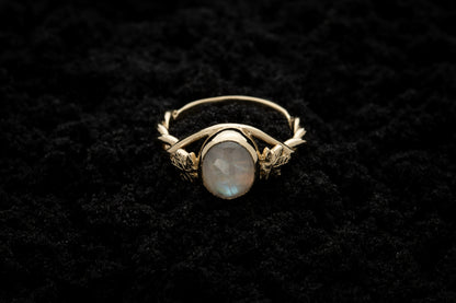 A botanical engagement ring made from rainbow moonstone and 9 carat yellow gold lays atop black sand. It features a twisted band and an ivy leaf on each shoulder. The moonstone shines gently in the camera flash, teasing it's bluey rainbow hues.