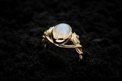 A botanical engagement ring made from rainbow moonstone and yellow gold sits, half submerged, in black sand. It features a twisted band and an ivy leaf on each shoulder. The moonstone shines gently in the camera flash, teasing it's bluey rainbow hues.