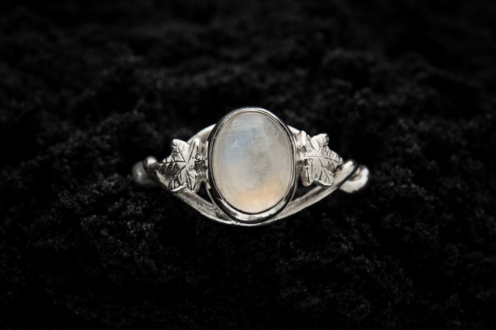  A botanical engagement ring made from rainbow moonstone and silver sits in black sand. It is photographed from above and features a twisted band and an ivy leaf on each shoulder. The moonstone shines gently in the camera flash, teasing it's bluey rainbow hues.