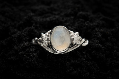 A botanical engagement ring made from rainbow moonstone and silver sits in black sand. It is photographed from above and features a twisted band and an ivy leaf on each shoulder. The moonstone shines gently in the camera flash, teasing it's bluey rainbow hues.