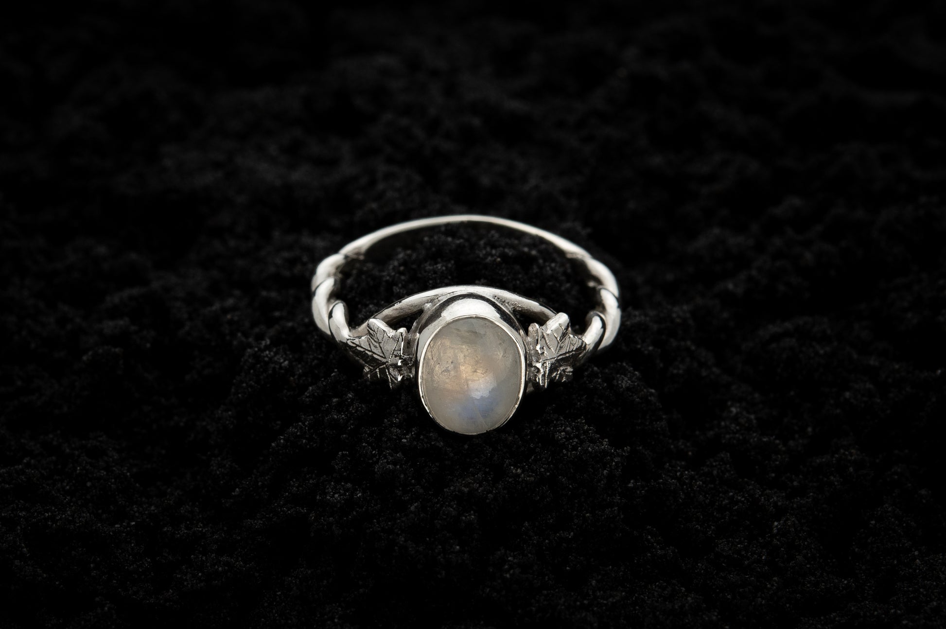 A botanical engagement ring made from rainbow moonstone and 9 carat white gold lays atop black sand. It features a twisted band and an ivy leaf on each shoulder. The moonstone shines gently in the camera flash, teasing it's bluey rainbow hues.