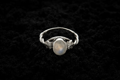 A botanical engagement ring made from rainbow moonstone and 9 carat white gold lays atop black sand. It features a twisted band and an ivy leaf on each shoulder. The moonstone shines gently in the camera flash, teasing it's bluey rainbow hues.