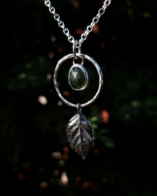 A silver pendant hangs from a chain in front of dark green, out-of-focus background. It is comprised of a rose-cut, green rutile quartz bezel-set in silver and surrounded by a hammered silver halo-like circlet. A solid silver bramble leaf hangs rom the bottom of the circlet. Everything is attached by tiny jump rings so it can move naturally. The leaf is oxidised and polished to reveal the natural details including veins. The pendant is illuminated in natural sunlight.