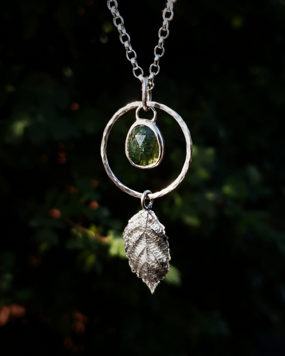 A silver pendant hangs from a chain in front of dark green, out-of-focus background. It is comprised of a rose-cut, green rutile quartz bezel-set in silver and surrounded by a hammered silver halo-like circlet. A solid silver bramble leaf hangs rom the bottom of the circlet. Everything is attached by tiny jump rings so it can move naturally. The leaf is oxidised and polished to reveal the natural details including veins. The pendant is illuminated in natural sunlight.