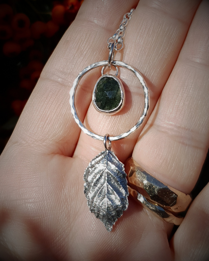 A silver pendant lays on an open palm to demonstrate its size. It is comprised of a rose-cut, green rutile quartz bezel-set in silver and surrounded by a hammered silver halo-like circlet. A solid silver bramble leaf hangs rom the bottom of the circlet. Everything is attached by tiny jump rings so it can move naturally. The leaf is oxidised and polished to reveal the natural details including veins. The pendant is illuminated in natural sunlight.
