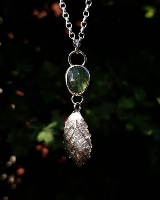A silver pendant hangs from a chain in front of dark green, out-of-focus background. It is comprised of a rose-cut, green rutile quartz bezel-set in silver. Below it hangs a solid silver bramble leaf, roughly twice the size of the gemstone, attached by tiny jump rings so it can move freely. The leaf is complete with natural details including veins. The pendant is illuminated in natural sunlight.