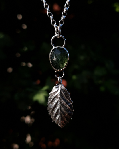 A silver pendant hangs from a chain in front of dark green, out-of-focus background. It is comprised of a rose-cut, green rutile quartz bezel-set in silver. Below it hangs a solid silver bramble leaf, roughly twice the size of the gemstone, attached by tiny jump rings so it can move freely. The leaf is complete with natural details including veins. The pendant is illuminated in natural sunlight.