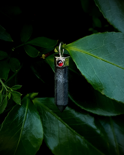 A black tourmaline point hangs from a branch surrounded by lush green leaves and foliage. The tourmaline is matte in contrast to the juicy red garnet and shiny silver. The image is lit by natural daylight.