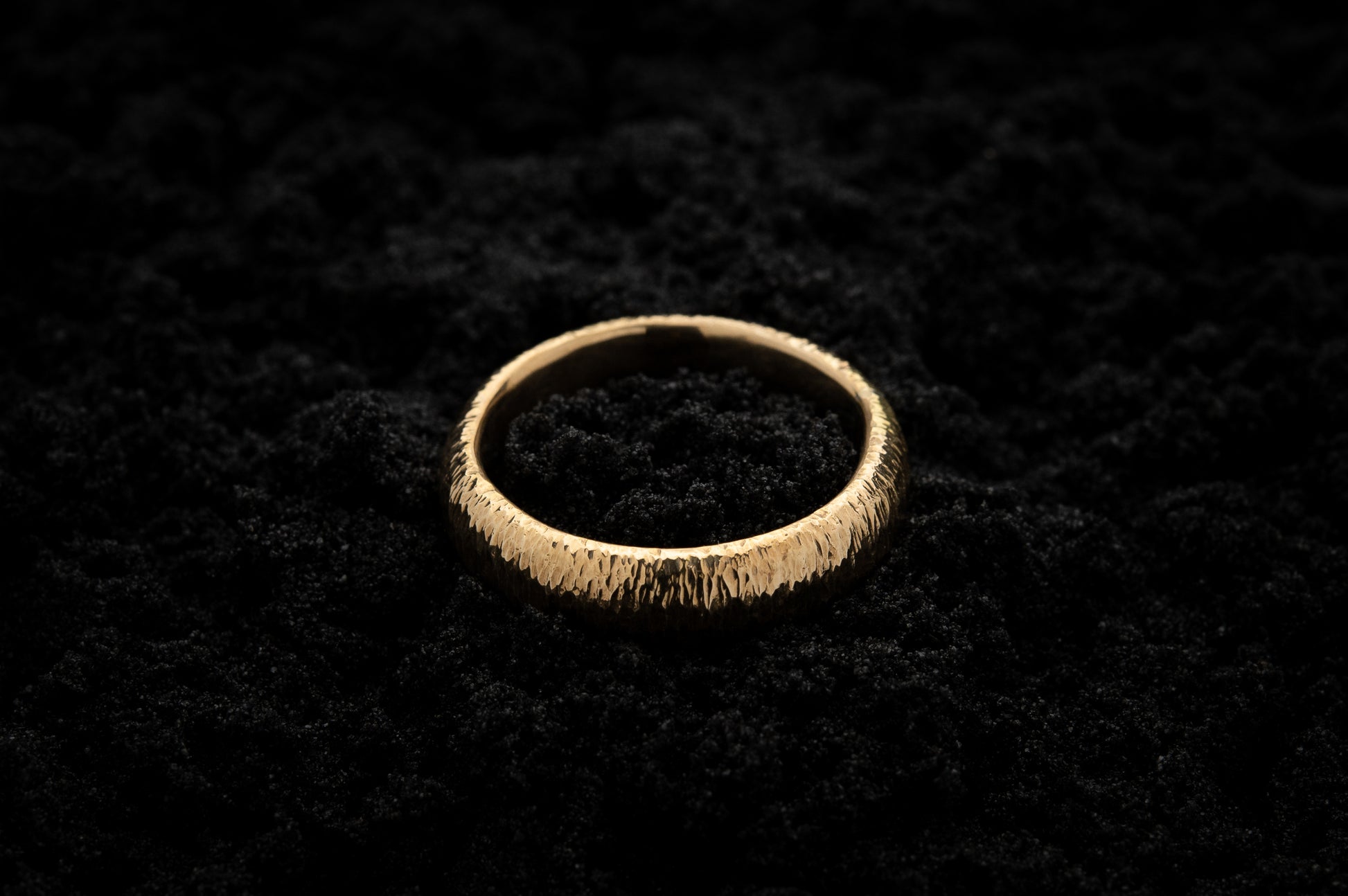 A thick yellow gold wedding ring sits in black sand to contrast the warm luscious glow. There are hammered striations around the band like the suns rays.