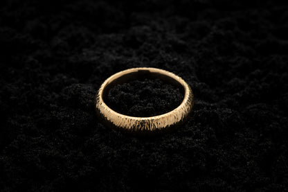A thick yellow gold wedding ring sits in black sand to contrast the warm luscious glow. There are hammered striations around the band like the suns rays.