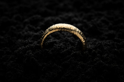 A thick yellow gold wedding ring sits in black sand to contrast the warm luscious glow. There are hammered striations around the band like the suns rays. It is half submerged in the sand and breaks through the surface like a golden rainbow.