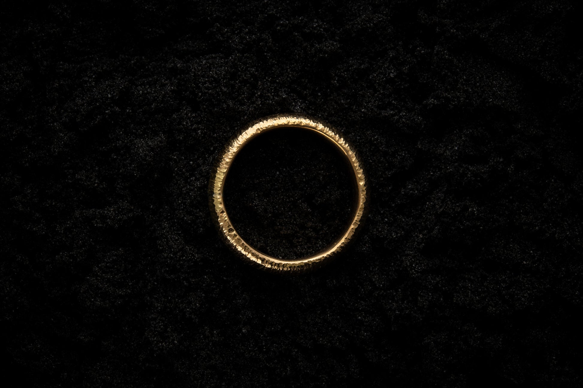 A thick yellow gold wedding ring sits in black sand to contrast the bright, warm, luscious glow. There are hammered striations around the band like the suns rays. It is photographed from above.