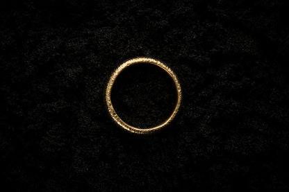 A thick yellow gold wedding ring sits in black sand to contrast the bright, warm, luscious glow. There are hammered striations around the band like the suns rays. It is photographed from above.