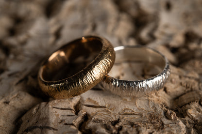 A yellow and white gold matching wedding band pair sit a wooden dried bark surface. The rings have hammered striations that glisten in the light. The inside of the bands are smooth and look comfortable.