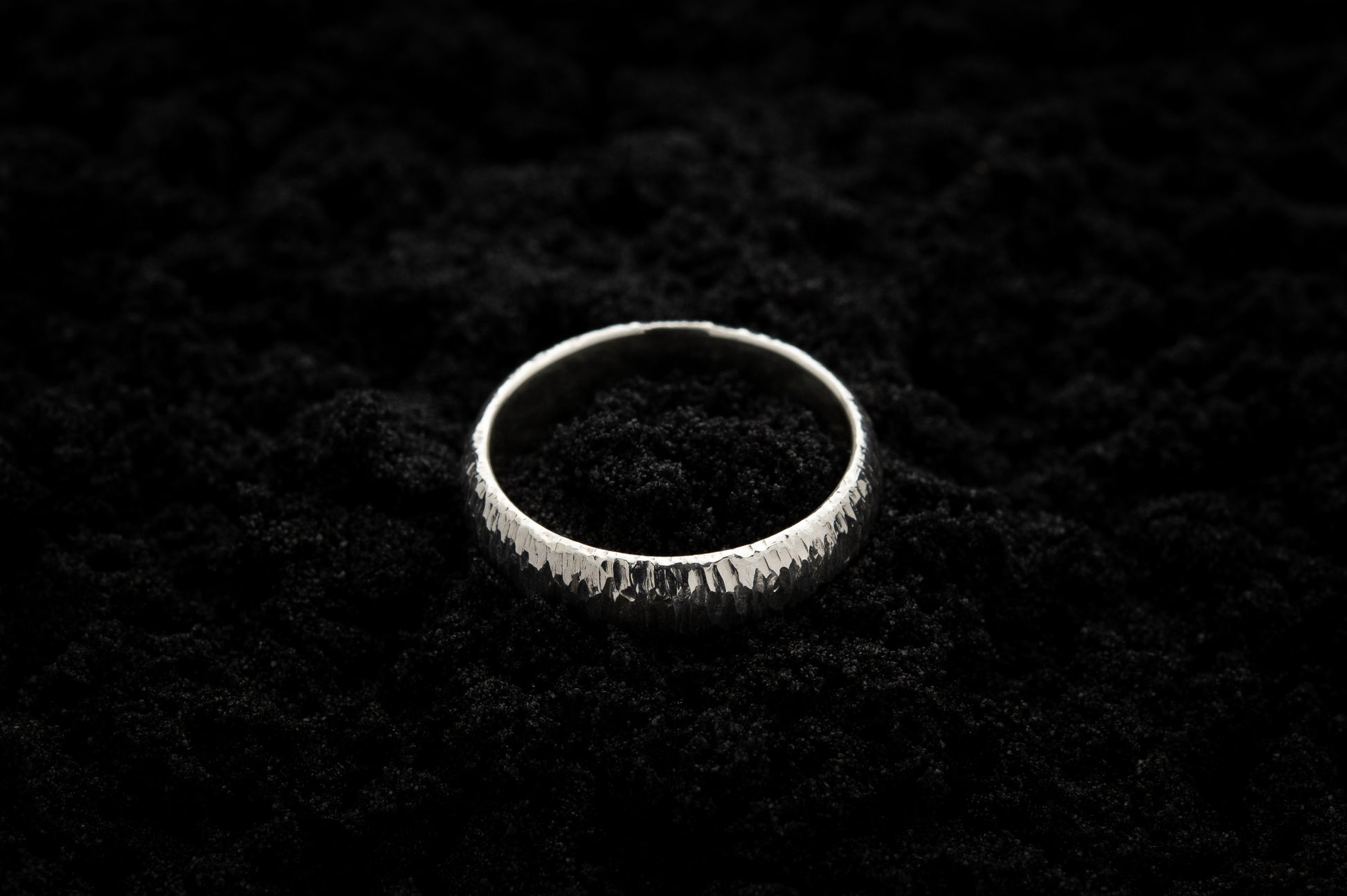 A thick white gold ring sits in black sand to contrast the bright luscious glow. There are hammered striations around the band like the suns rays.