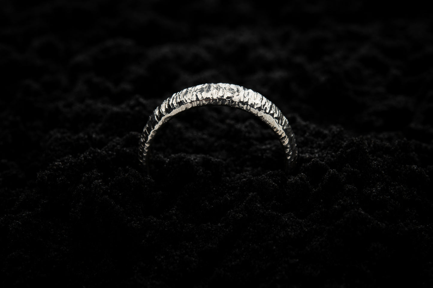 A thick white gold wedding ring sits in black sand to contrast the bright lux glow. There are hammered striations around the band like the suns rays. It is half submerged in the sand and breaks through the surface like a silver rainbow.