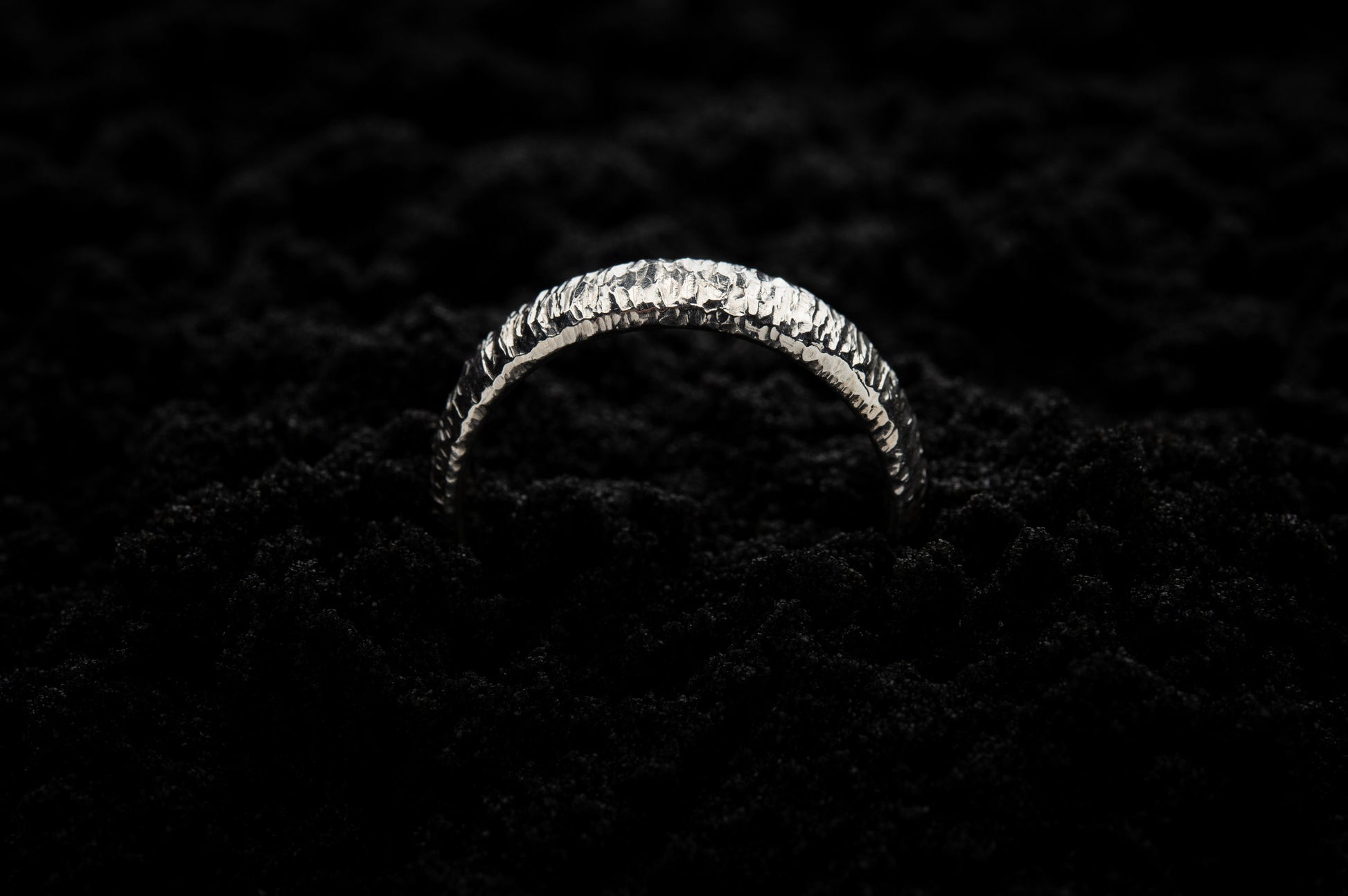 A thick white gold wedding ring sits in black sand to contrast the bright lux glow. There are hammered striations around the band like the suns rays. It is half submerged in the sand and breaks through the surface like a silver rainbow.
