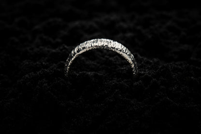A thick white gold wedding ring sits in black sand to contrast the bright lux glow. There are hammered striations around the band like the suns rays. It is half submerged in the sand and breaks through the surface like a silver rainbow.