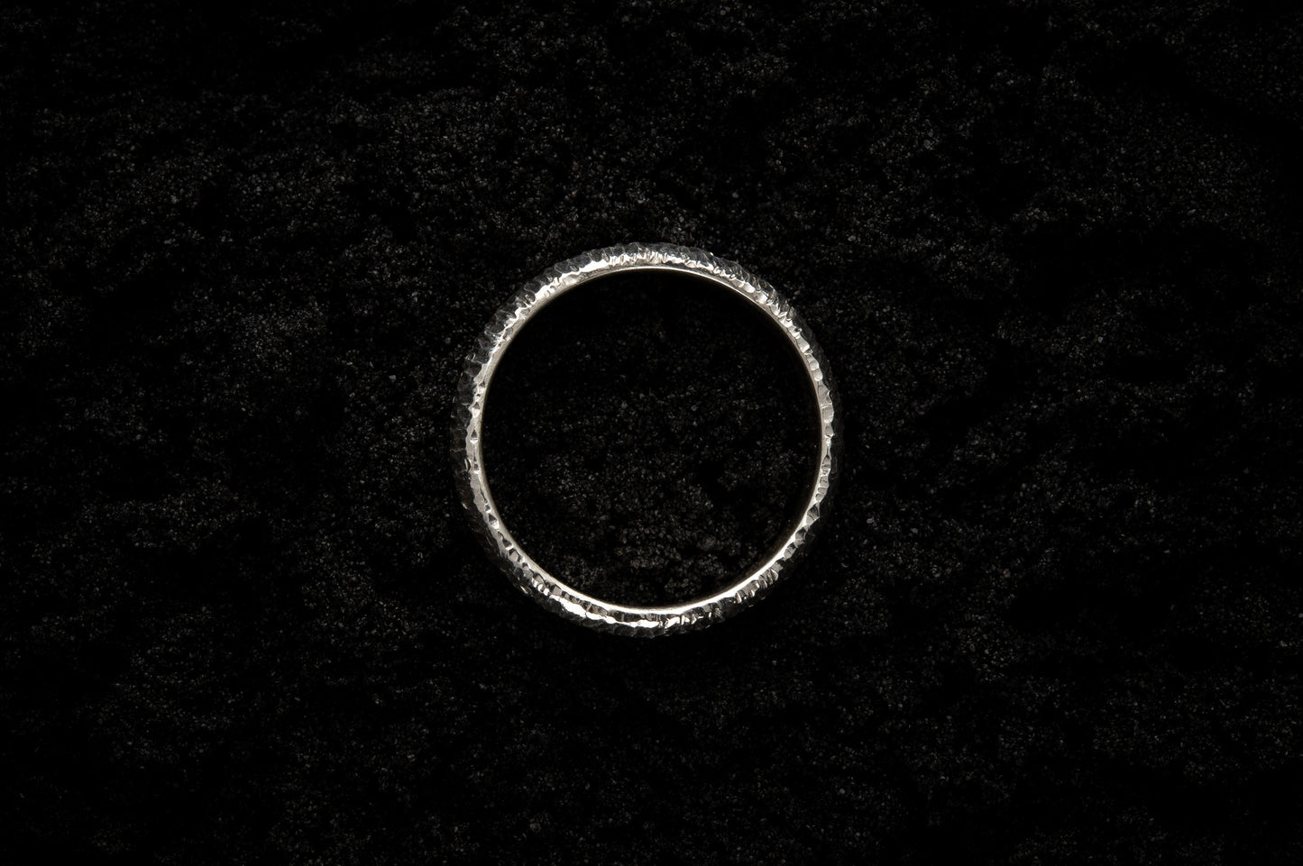 A thick silver wedding ring sits in black sand to contrast the bright luscious shine. There are hammered striations around the band like the suns rays.