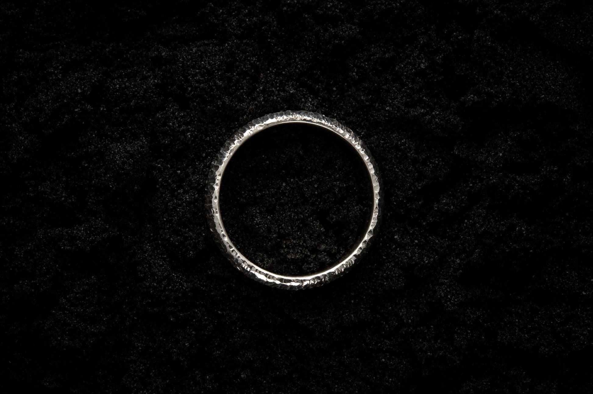 A thick silver wedding ring sits in black sand to contrast the bright luscious shine. There are hammered striations around the band like the suns rays.