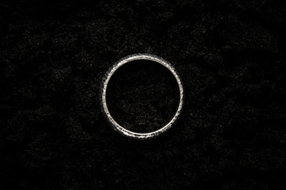 A thick silver wedding ring sits in black sand to contrast the bright luscious shine. There are hammered striations around the band like the suns rays.