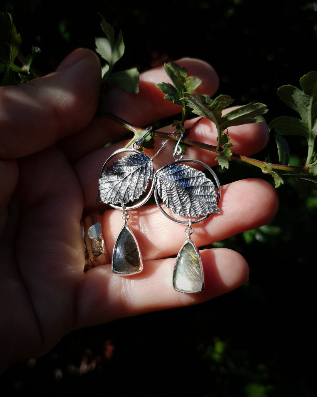 Two silver earrings are held in an open palm to demonstrate their size. They are comprised of a silver circlet with a solid silver bramble leaf in front of it. A small teardrop shaped labradorite hangs below it from the circlet. The labradorites shine turquoise and golden-brown in the light; and the bramble leaves are complete with natural details including veins. The earrings are illuminated in natural sunlight.