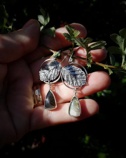 Two silver earrings are held in an open palm to demonstrate their size. They are comprised of a silver circlet with a solid silver bramble leaf in front of it. A small teardrop shaped labradorite hangs below it from the circlet. The labradorites shine turquoise and golden-brown in the light; and the bramble leaves are complete with natural details including veins. The earrings are illuminated in natural sunlight.