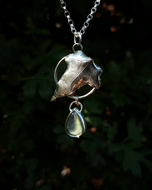 Silver pendant comprised of a silver circlet from which a solid silver ivy leaf hangs from a chain. A small teardrop shaped labradorite hangs below it from the circlet. The labradorite is shining dark turquoise and a golden-green in the light; and the ivy leaf is complete with natural details including veins. In the background is out of focus natural greenery and the pendant is bathed in natural sunlight.