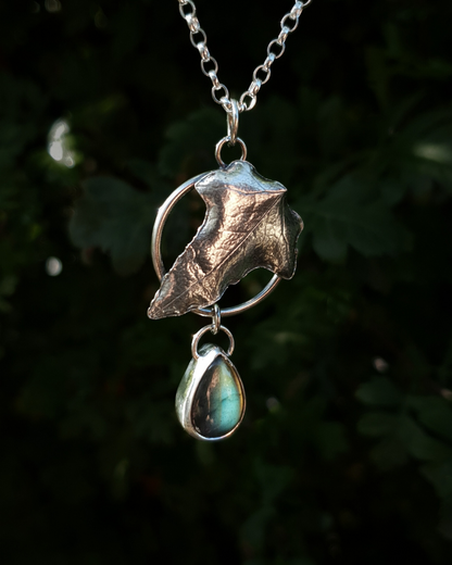 Silver pendant comprised of a silver circlet from which a solid silver ivy leaf hangs from a chain. A small teardrop shaped labradorite hangs below it from the circlet.  The labradorite is shining dark blue and a pale irridescent turquoise in the light; and the ivy leaf is complete with natural details including veins. In the background is out of focus natural greenery and the pendant is bathed in natural sunlight.