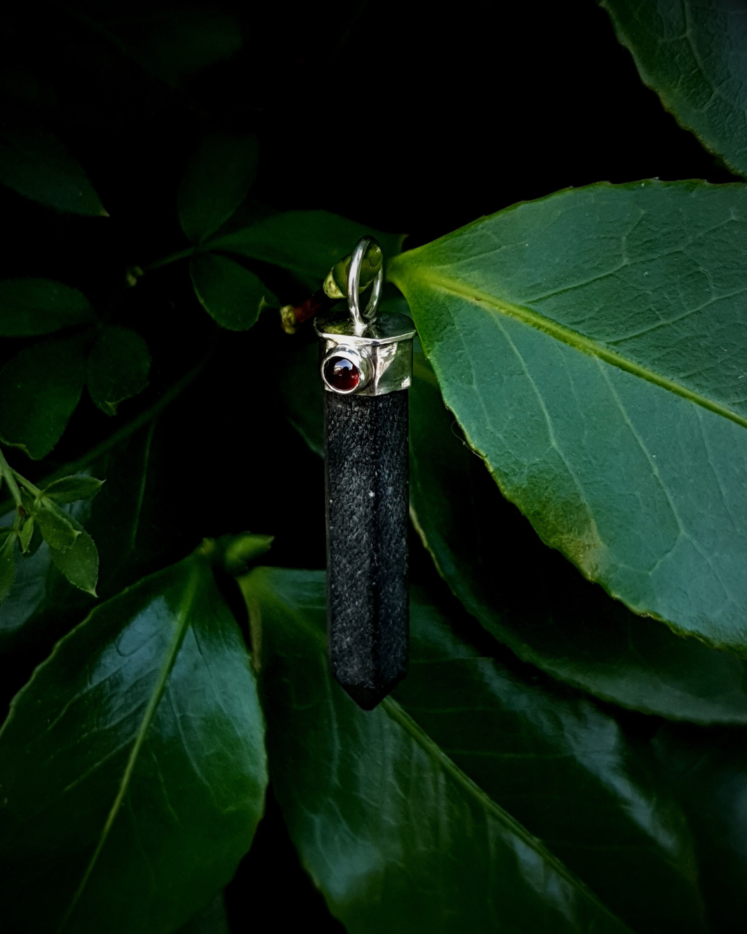 A black tourmaline point hangs from a branch surrounded by lush green leaves and foliage. The tourmaline is matte in contrast to the juicy red garnet and shiny silver. The image is lit by natural daylight.
