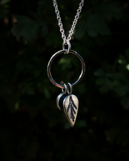 Silver pendant comprised of a silver circlet from which a solid silver upside-down teardrop, imprinted with a cypress sprig, and moss agate pendant hang from a silver chain. The moss agate is a small oval with dark green and white details; and the teardrop charm is approximately twice it's length. In the background is out of focus natural greenery and the charms are illuminated in natural sunlight.
