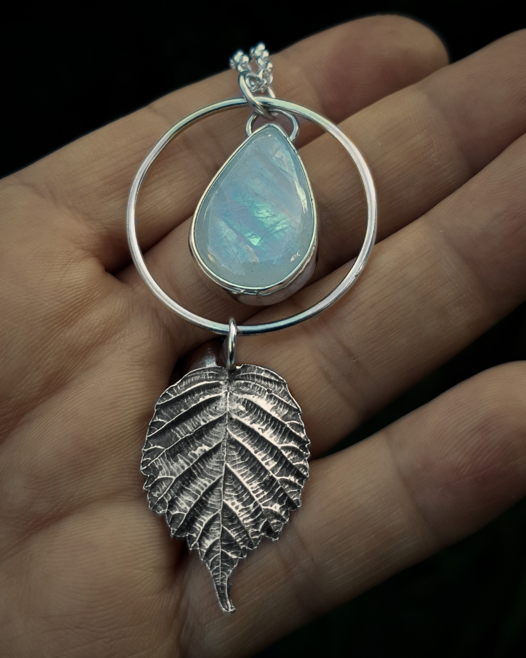 A silver pendant lays in an open palm to demonstrate its size. It is comprised of a large teardrop-shaped rainbow moonstone bezel-set in silver and surrounded by a silver halo-like circlet. A solid silver bramble leaf hangs rom the bottom of the circlet. Everything is attached by tiny jump rings so it can move naturally. The leaf is oxidised and polished to reveal the natural details including veins. The pendant is illuminated in natural sunlight showing the iridescent play of rainbows in the moonstone.
