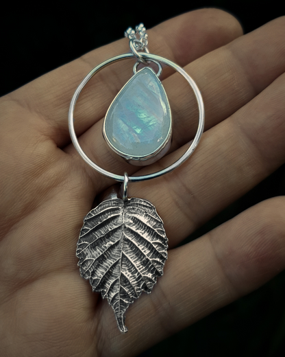 A silver pendant lays in an open palm to demonstrate its size. It is comprised of a large teardrop-shaped rainbow moonstone bezel-set in silver and surrounded by a silver halo-like circlet. A solid silver bramble leaf hangs rom the bottom of the circlet. Everything is attached by tiny jump rings so it can move naturally. The leaf is oxidised and polished to reveal the natural details including veins. The pendant is illuminated in natural sunlight showing the iridescent play of rainbows in the moonstone.