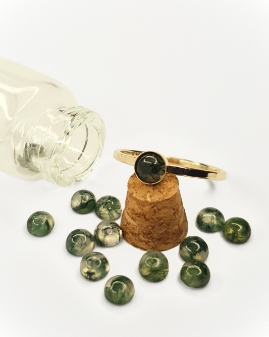 A little, glass bottle lays on its side with its content spilled out over a white background. The contents are 12 little moss agate gemstones. A beautiful, delicate, 9 carat gold ring with a moss agate rests against the cork stopper of the class bottle.