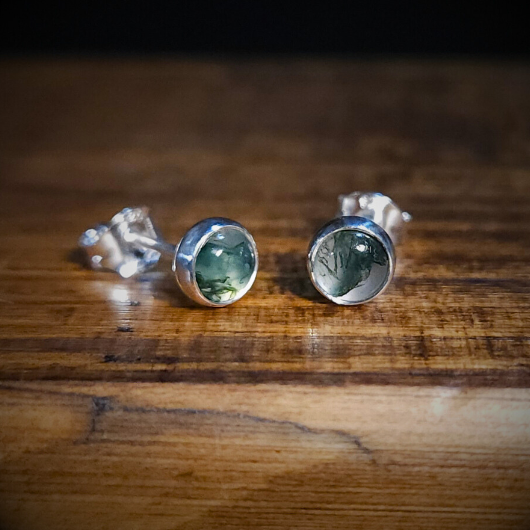 A pair of round, moss agate & sterling silver stud earrings sit on a wooden surface.