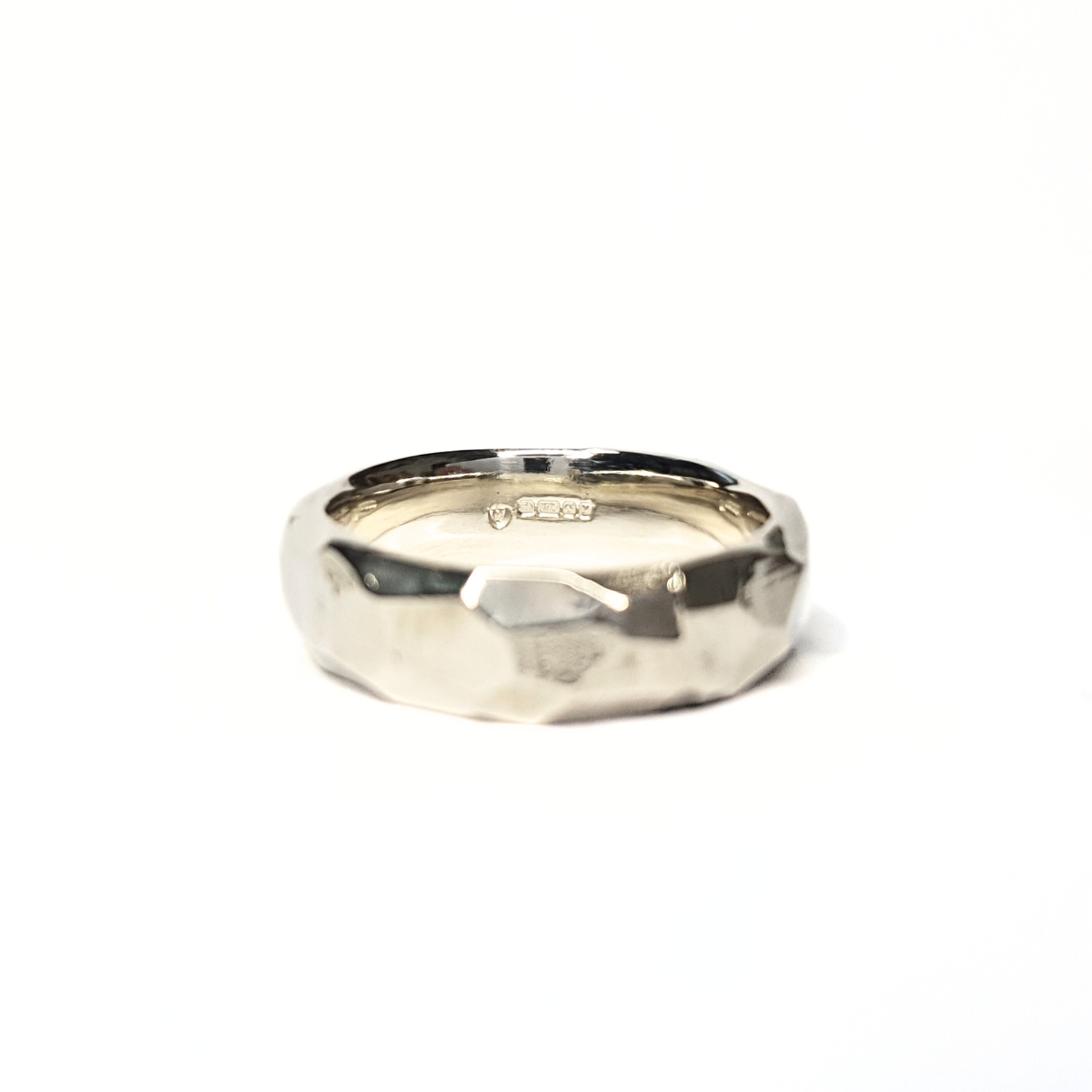 A 9ct white gold rough cut and faceted ring sits against a crisp, white background. The front of the ring is out of focus but the inside of the ring is in focus showing the hallmarks.