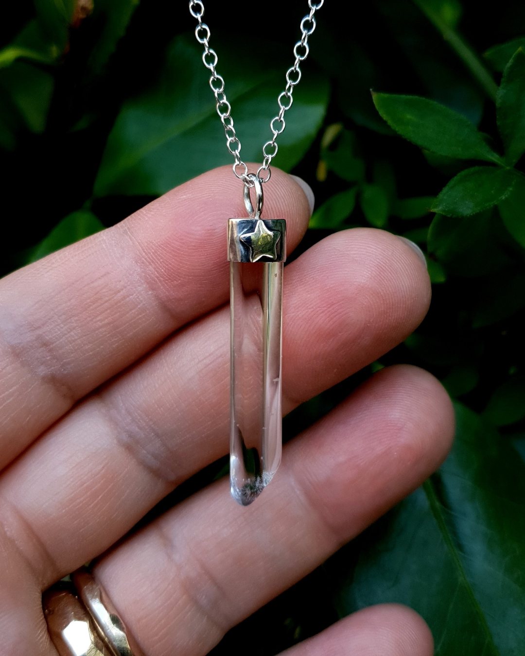 A pendant hangs from a dark green bush, propped up by the photographers fingers to show the size of it. The pendant comprises of a clear quartz point in a sterling silver setting. A fir tree has been stamped into the silver setting and oxidised black so that the detail stands out. 