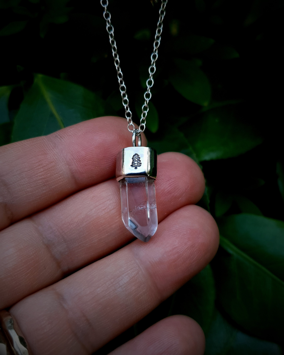 A pendant hangs from a dark green bush, propped up by the photographers fingers to show the size of it. The pendant comprises of a clear quartz point in a sterling silver setting. A fir tree has been stamped into the silver setting and oxidised black so that the detail stands out. 