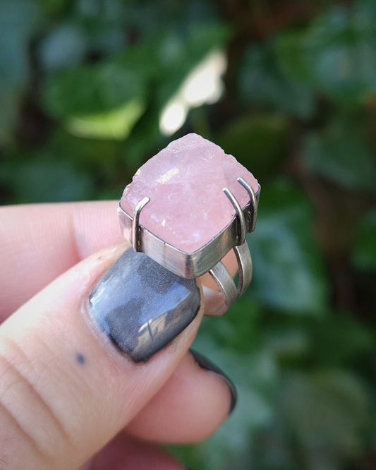A sterling silver Ring with a square rose quartz is held by a hand against a blurred green background of green foliage