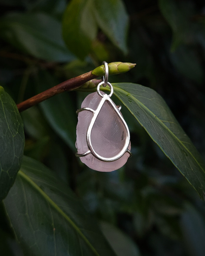 A pastel-pink, teardrop-shaped rose quartz pendant hangs from a thin, leafy branch. It's photographed from the back, hanging amongst dark leafy foliage, to show it's minimal sterling silver setting - designed to allow the light through the stone and highlight it's rich texture and colour.