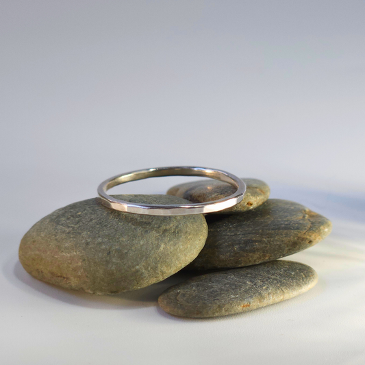 A slim, hammered, sterling silver ring sits on a little pile of beach pebbles against a neutral backdrop. The light, coming in from the right, shines gorgeously, reflecting in it's smooth, hammered edges.