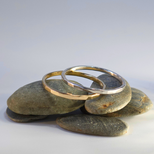 A hammered, dainty, sterling silver and 9ct yellow gold sit on a pile of pebbles on a light, neutral backdrop. Their smooth, shiny, hammered edges reflect the light beautifully.