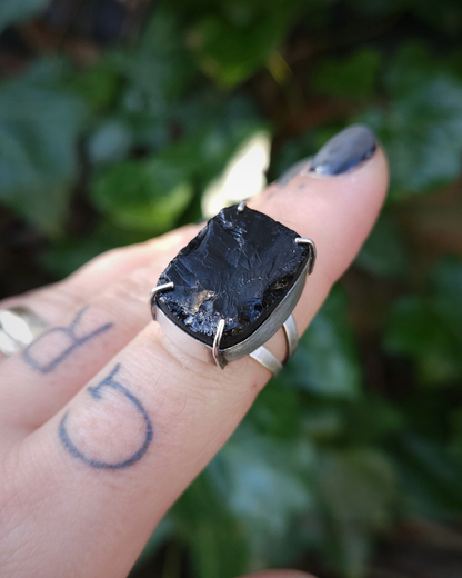 A sterling silvering ring with a rectangular smoky quartz is being held on the end of a finger against a botanical background, which is out of focus. The sterling silver is recycled and has a softly brushed finish and the smoky quartz is dark and full of texture as it is roughly cut and raw.