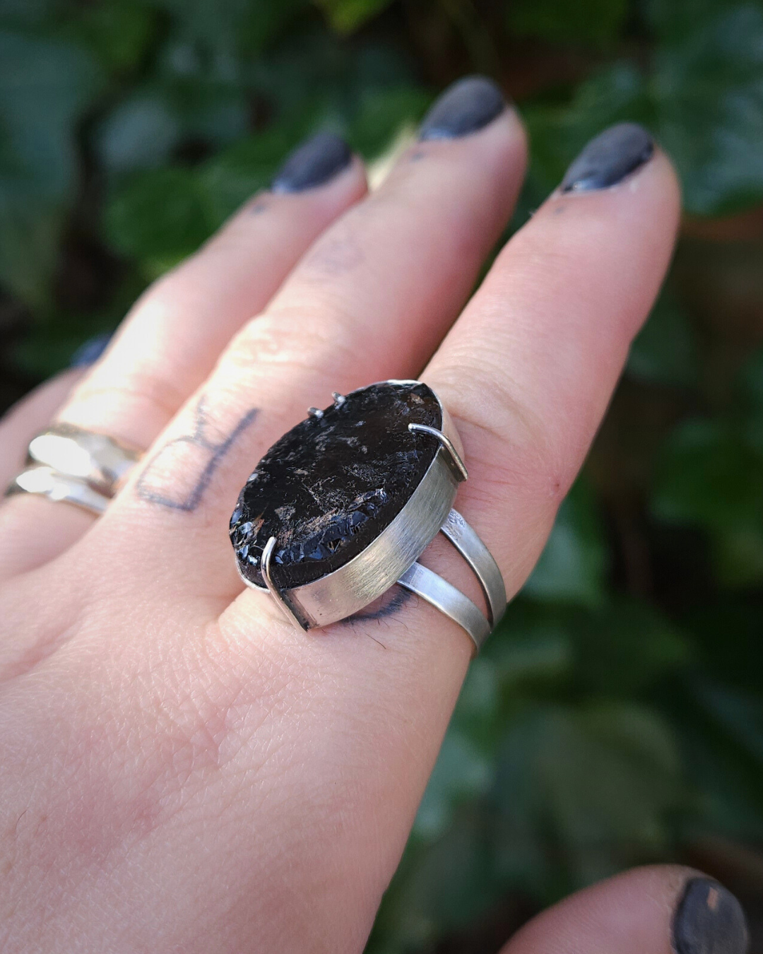 A hand wears a gothic style smoky quartz and silver ring. The fingers have tattoos and black nails and the hand is held up against a green leafy background.