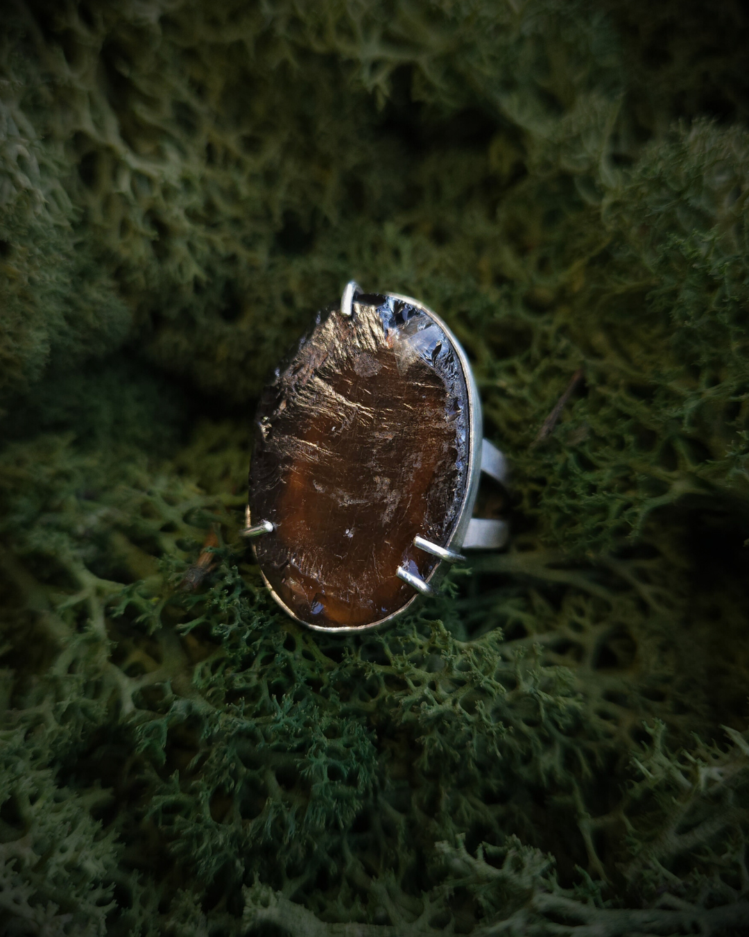 A claw-set smoky quartz and silver ring rests in a bed of green moss. It's androgynous in style with a modern setting but a natural, rough-cut stone.
